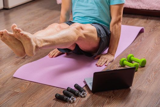 50-year-old man performs exercises while lying on the rug at home, looking at the computer. During a pandemic, a person trains in an apartment via the Internet.