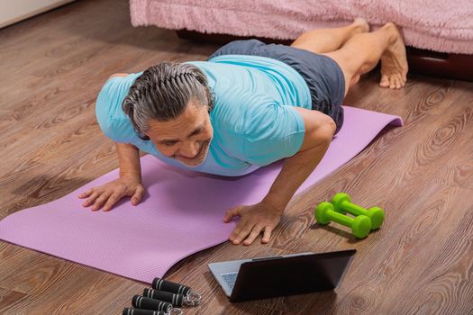 50-year-old man performs exercises while lying on the rug at home, looking at the computer. During a pandemic, a person trains in an apartment via the Internet.