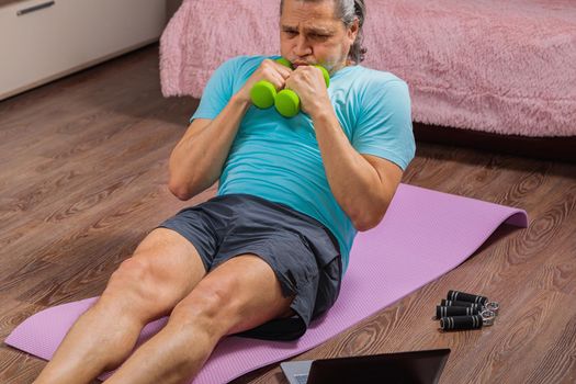 50-year-old man performs exercises while lying on the rug at home, looking at the computer. During a pandemic, a person trains in an apartment via the Internet.