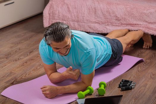 50-year-old man performs exercises while lying on the rug at home, looking at the computer. During a pandemic, a person trains in an apartment via the Internet.