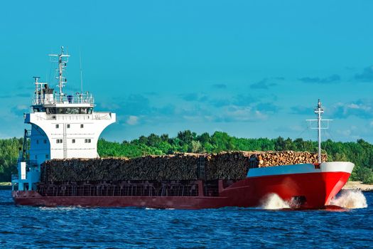 Red cargo ship fully loaded with wood moving at clear day