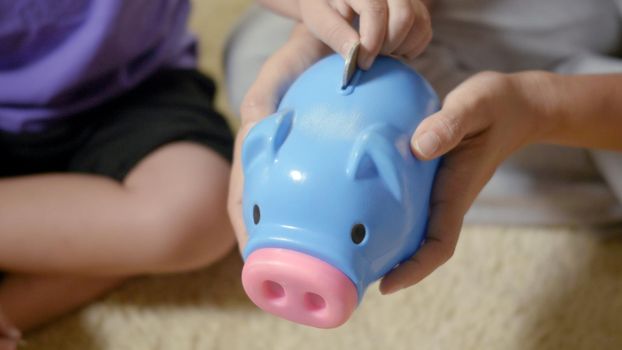 Happy Asian kid boy preschool and mother putting pin money coins into piglet. Little child putting coin into piggy bank for saving with pile of coins at home, Investment education concept,