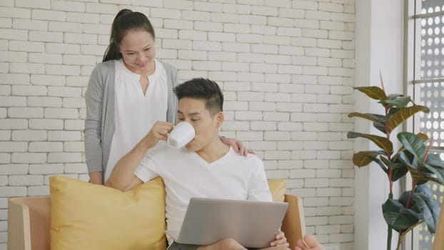 Happy Asian beautiful family couple husband and wife laughing sitting on sofa in the living room working with laptop computer at home. Woman brings coffee to the man during work