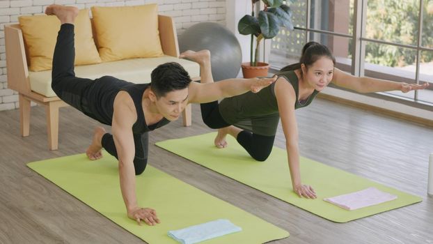 Happy Asian beautiful lifestyle family couple doing YOGA standing meditating on virabhadrasana 3 pose workout at home together on mat. Two people sport healthy concept