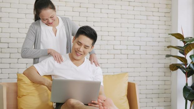 Happy Asian family couple husband and wife laughing sitting on sofa using laptop computer working from home. Business man sitting on sofa home working on laptop her woman is giving her shoulder massage