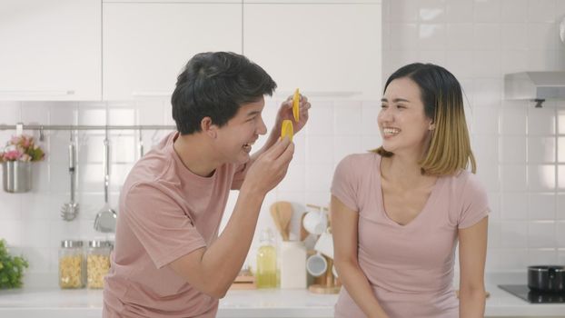 Happy Asian beautiful young family couple husband and wife enjoying smile and laugh holds a cut orange in front of the guy's eyes spending time together in kitchen at home.