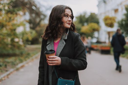 Calm young female holding a paper cup outdoors while standing in the street and looking into the distance