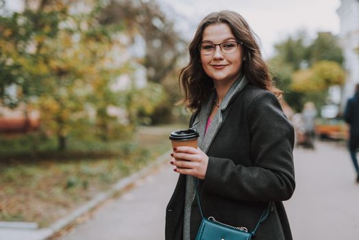 Gladsome bespectacled young lady holding a paper cup of coffee and smiling while standing in the street