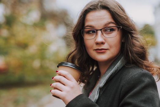 Curious bespectacled young woman looking into the distance while being outdoors with a paper cup of coffee in her hand