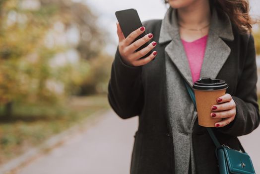 Cropped photo of a woman in a jacket holding a paper cup of coffee and a smartphone while standing outdoors