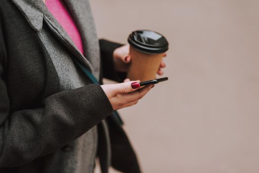 Cropped photo of a lady outdoors having a paper cup of coffee to go and a modern smartphone in her hands