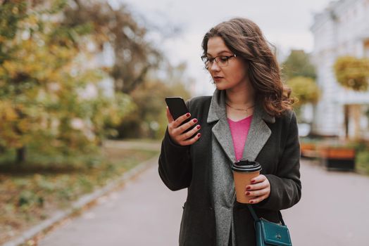 Serious young woman holding a carton cup of coffee while standing in the street with a smartphone and looking at the screen of it