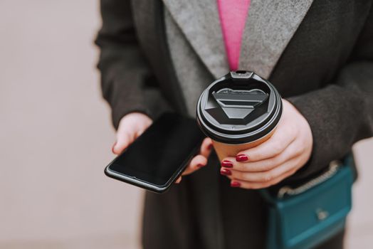 Close up photo of a lady with red manicure holding a modern smartphone and a paper cup of coffee