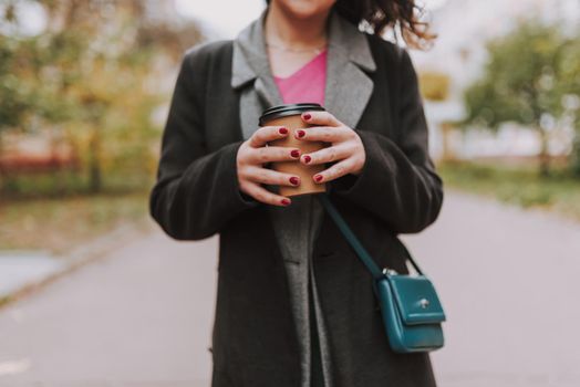 Cropped photo outdoors. Hands of woman with red manicure holding a paper cup of coffee