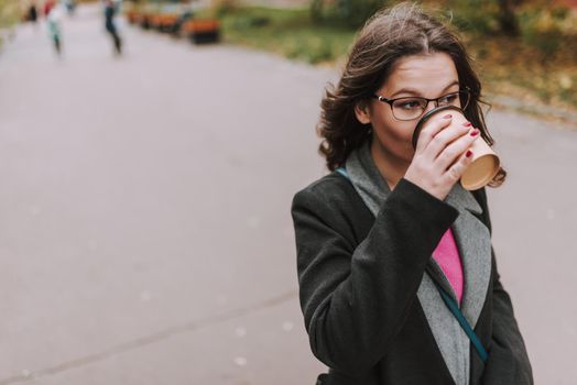 Bespectacled attractive young woman standing outdoors in a coat and looking into the distance while drinking coffee