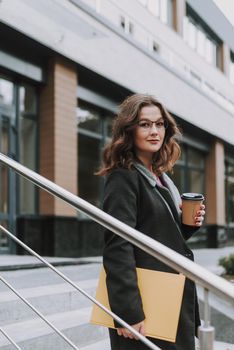 Confident pretty woman holding a book while standing with a paper cup of coffee on the stairs of a building