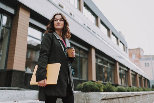 Beautiful lady in a coat holding a book and having a cup of coffee to go while standing outdoors and looking into the distance