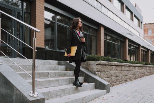 Pretty bespectacled young female having coffee outdoors and looking away while standing on the stairs with a book