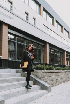 Pretty young woman standing on the stairs outside the building with a book and a paper cup of coffee and kindly smiling