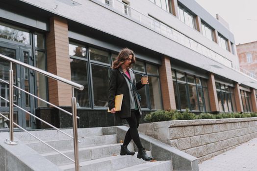 Beautiful young woman with a big book and a cup of coffee to go looking at her feet while walking downstairs