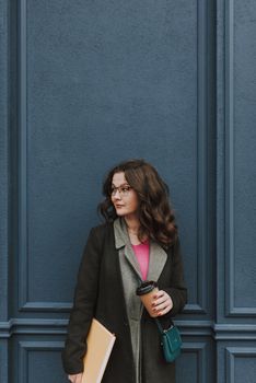 Calm young woman in spring clothes holding a book and a paper cup of coffee while standing by the dark blue wall