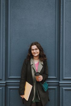 Positive lovely lady with a book smiling while standing by the dark blue wall and having a cup of coffee to go