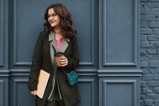 Cheerful bespectacled young woman with wavy hair holding a cup of coffee to go and a book and looking away with a smile