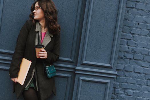 Calm attractive lady with red nails holding a paper cup of coffee and a big book while standing with her back to the blue wall. Website banner