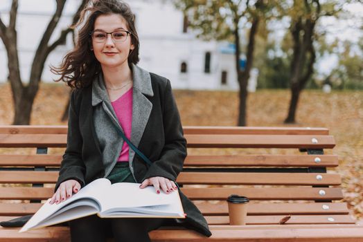 Smiling female student sitting on the bench with hot drink and book outdoors