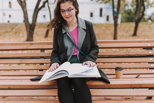 Pretty lady wearing glasses and looking at book while sitting on the bench in the park