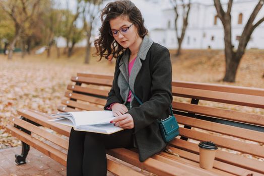 Attractive young lady wearing coat and glasses reading book while sitting on bench outdoors