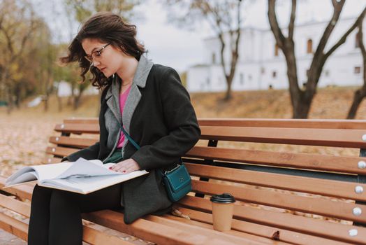 Beautiful female student wearing glasses and leafing through pages of book in a park while studying