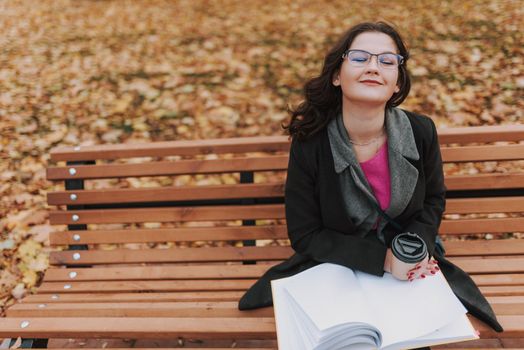 Top view of smiling attractive woman with eyes closed sitting on the bench and holding book, coffee in the park