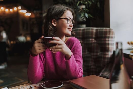 Waist up of smiling young woman sitting in coffee shop and looking away while holding cup