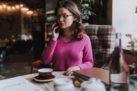 Waist up of attractive lady sitting at the table in coffee house and talking on smartphone