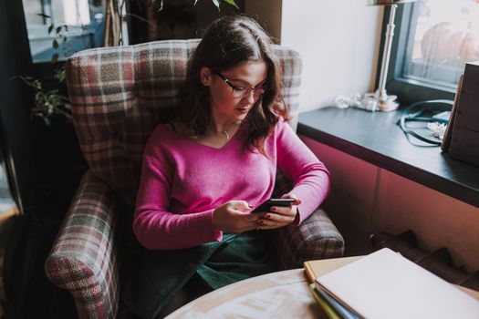 Pretty lady wearing glasses sitting in cafe while looking at smartphone screen and typing