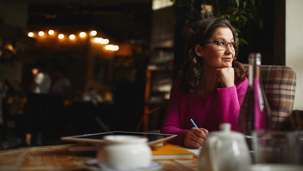 Smiling female wearing glasses and sitting in coffee house while making notes in notebook