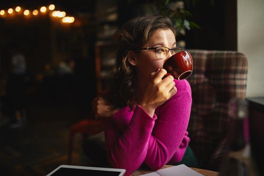 Pretty woman wearing glasses and sitting at the table in cafe while holding cup with hot drink