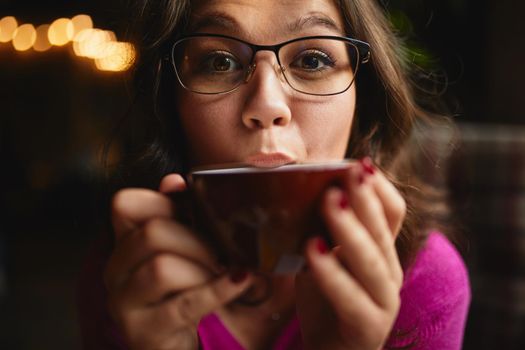 Close up of pretty young woman holding red cup od hot drink in cozy coffee house
