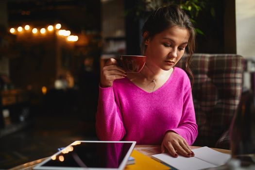 Beautiful brunette sitting in cozy cafe and studying while holding cup of coffee