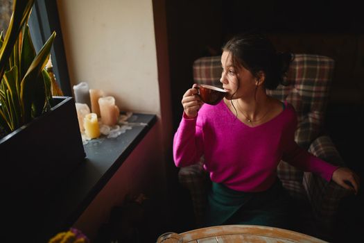 Top view of young female holding red cup and sitting in cafe while dreaming near window