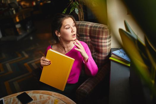 Attractive young brunette holding yellow notebook while spending her day off in cozy coffee house