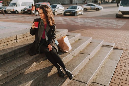 Happy pretty woman in coat sitting on steps while holding hot drink during sunny day