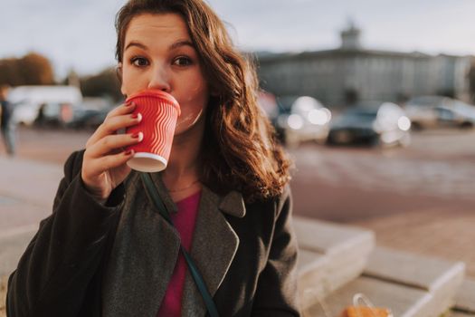 Close up of pretty young female standing in the city while enjoying coffee with cars on the background