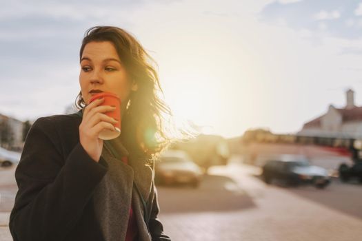 Waist up of attractive young woman standing on the street while holding coffee and looking away