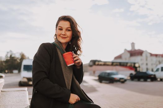 Waist up of smiling beautiful female sitting on steps while holding hot drink with car on the background