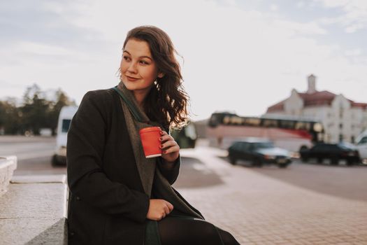 Happy pretty young lady posing with coffee while looking away and smiling