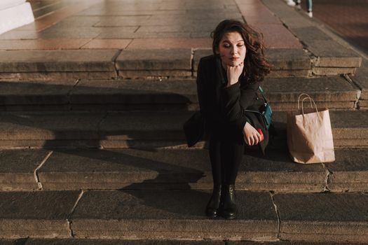 Smiling attractive lady sitting on steps with coffee and shopping bag next to her outdoors