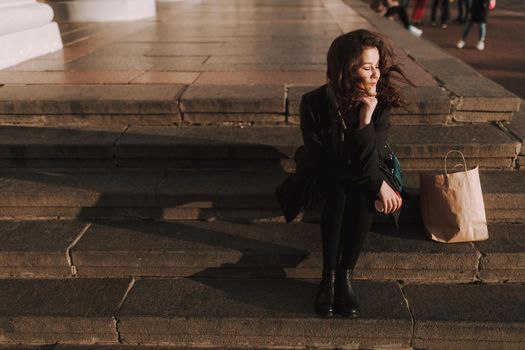 Pretty woman sitting on steps with hot drink and shopping bag next to her outdoors