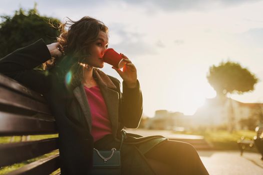 Happy attractive female sitting on bench while holding paper cup of hot drink outdoors
